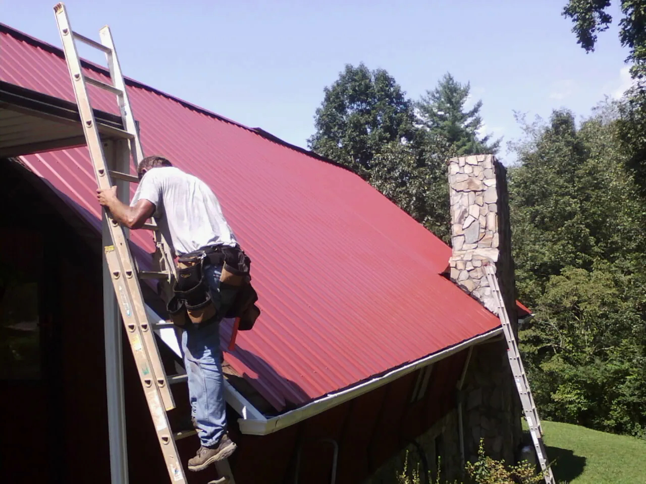 Roofer on a ladder beside a red metal roof and stone chimney — Soffit & Fascia Repair by Roofing Brothers in Englewood Cliffs